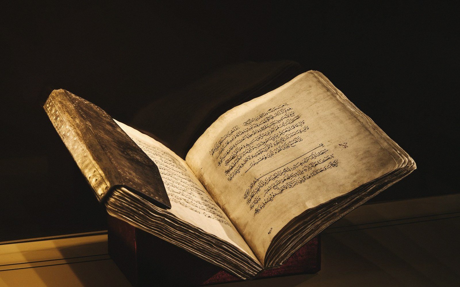 An old manuscript with Arabic script lies open on a stand under dim lighting